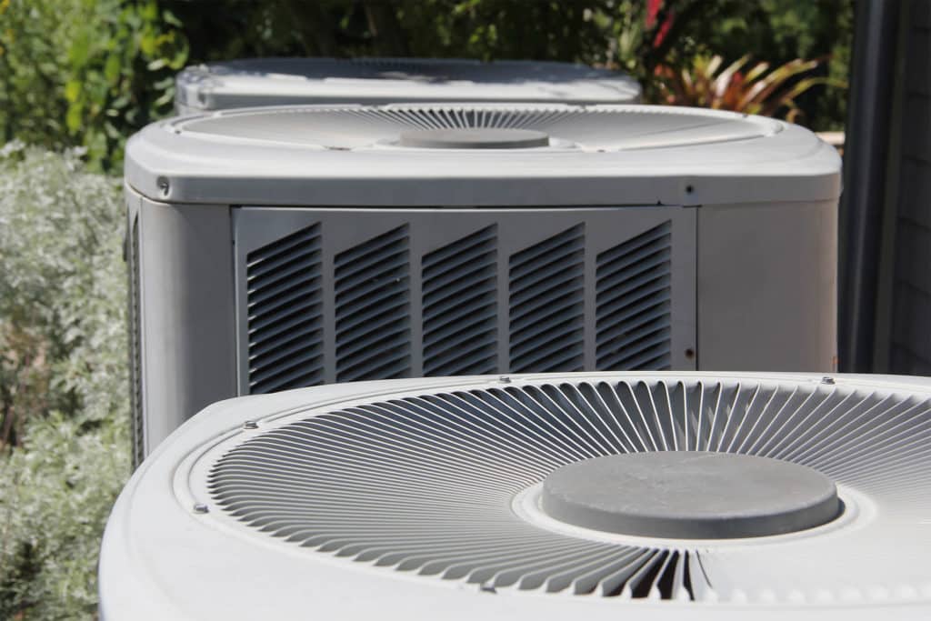 Two outdoor air conditioner condenser units running in bright sun beside a house