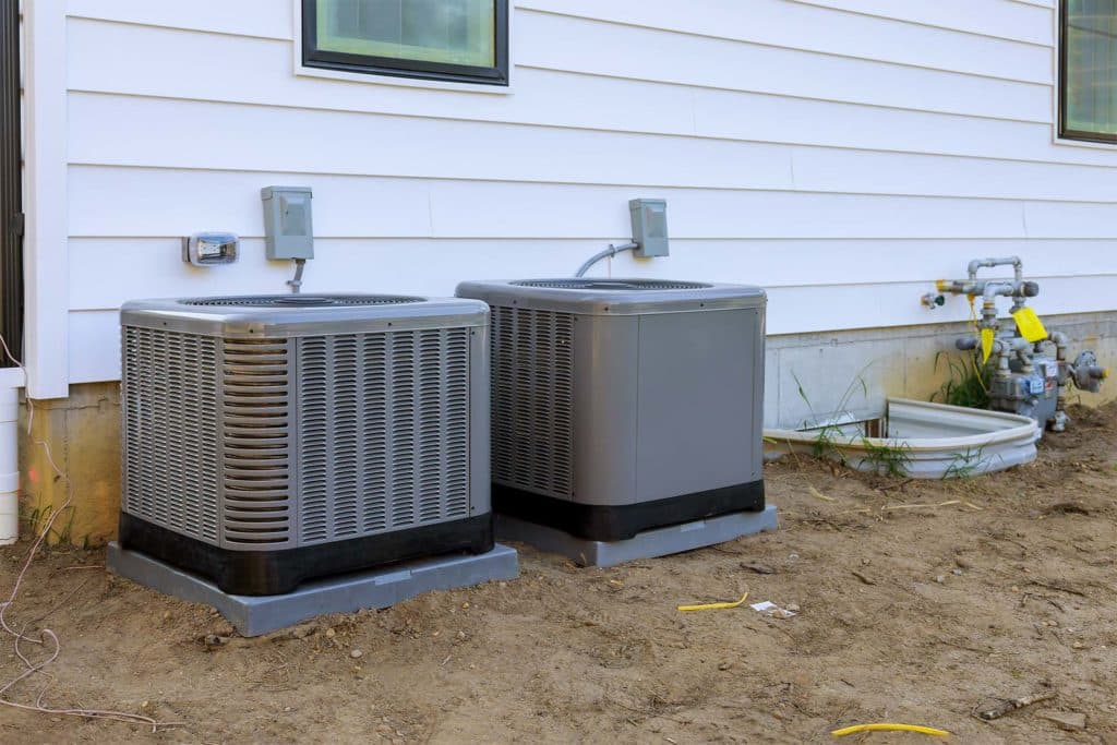 Two outdoor AC condenser units on pads beside a house, with bare ground and debris around them