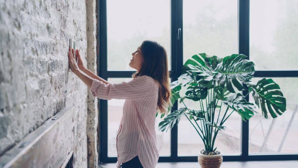 Woman adjusting a wall thermostat in a bright room with large windows and a potted plant